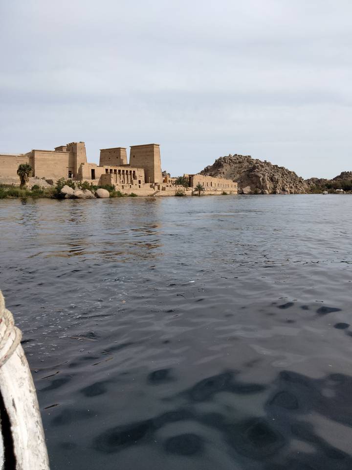 Ancient sandstone temple complex on an island beside the Nile River with rocky hill backdrop and rippling water in the foreground