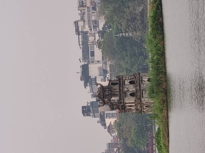 The historic Turtle Tower stands on a small grassy islet in Hoan Kiem Lake with Hanoi's skyline behind on a hazy day.