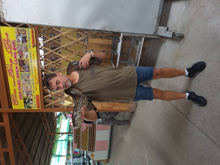 A tourist poses with a large python draped over his shoulders inside a rustic market building.