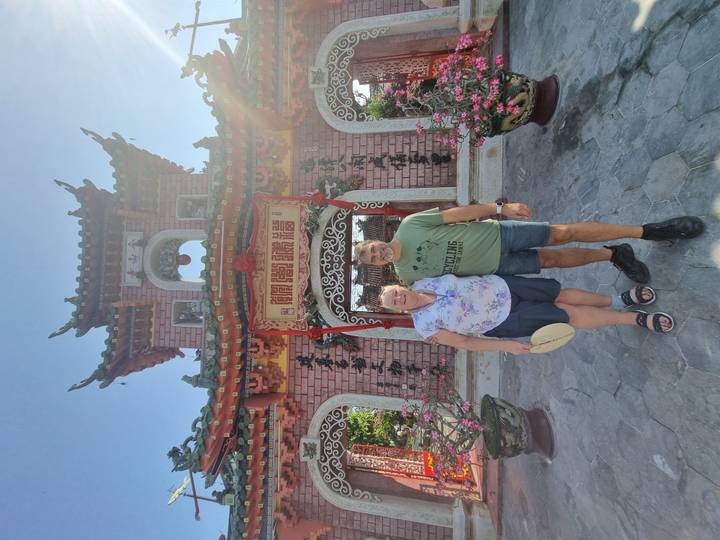 Smiling couple stand before the ornate gateway of the Phuoc Kien Assembly Hall in bright sunshine.