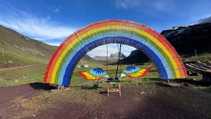 A vibrant sculptural rainbow arch frames the valley near Rainbow Mountain with snow-capped peaks beyond.