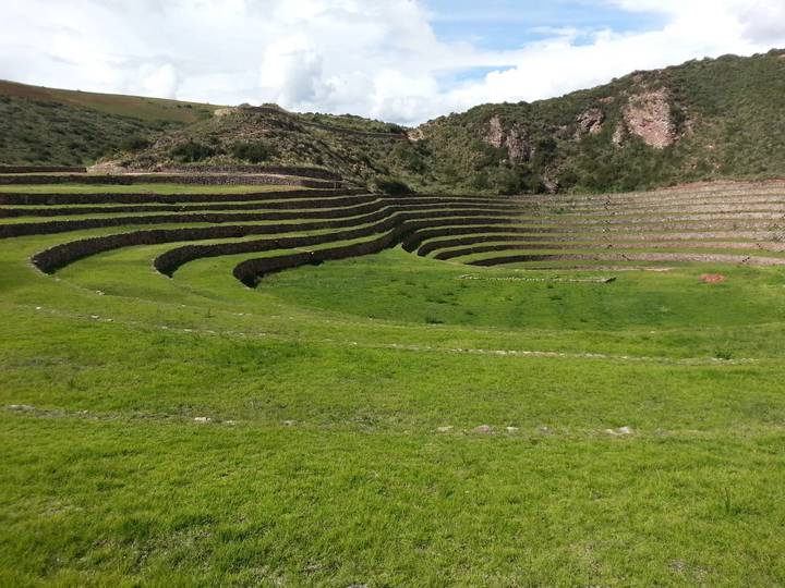 The concentric agricultural terraces of Moray curve through lush green grass under a partly cloudy sky.