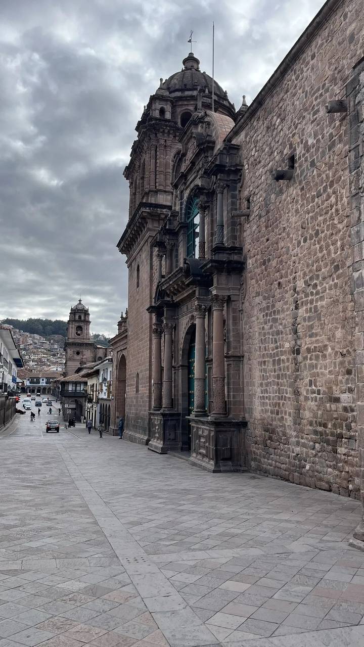 Stone façade of Cusco Cathedral lines a narrow street with the bell tower of Iglesia de la Compañía visible under a grey sky.