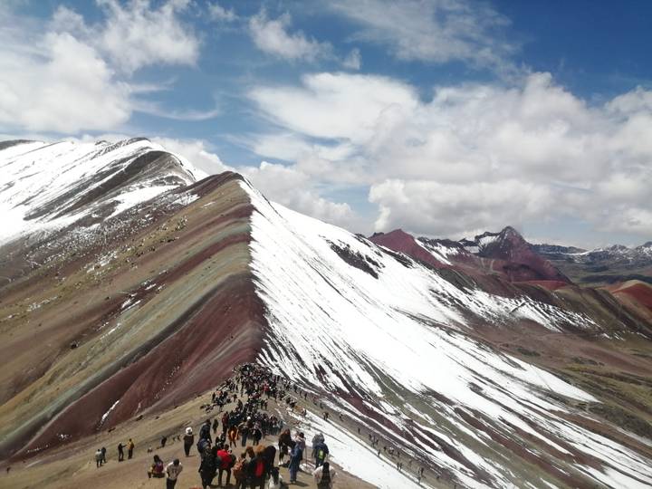 A dramatic ridge of Rainbow Mountain shows colorful striations beside snow-covered slopes with trekkers visible below.
