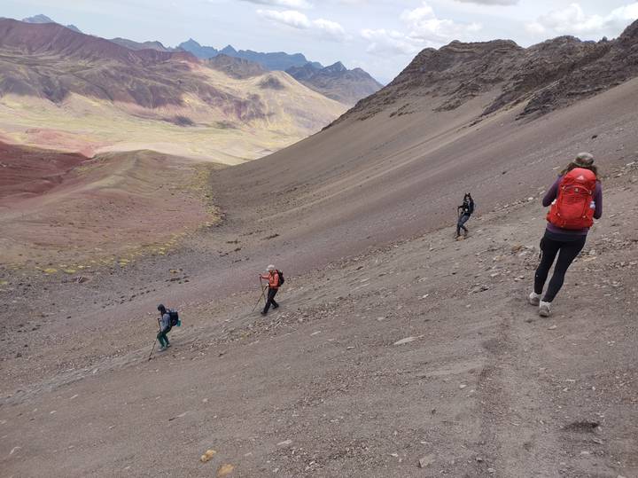 Hikers descend a steep scree slope with rainbow-tinted mountains and valleys stretching beyond.