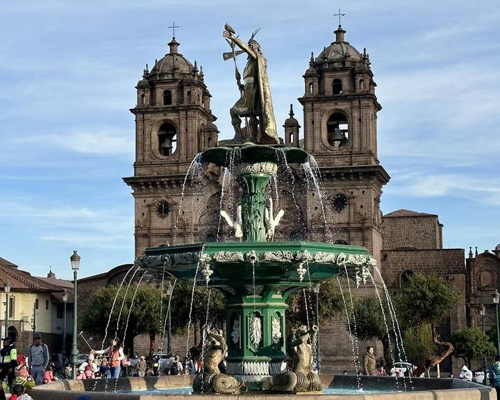 Ornate green fountain topped with an Incan figure splashes before the twin bell towers of Cusco's Iglesia La Compañía.