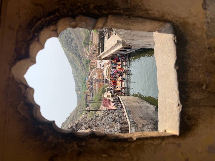 View through an ornate arched window to a sacred pool filled with pilgrims and surrounded by hills.