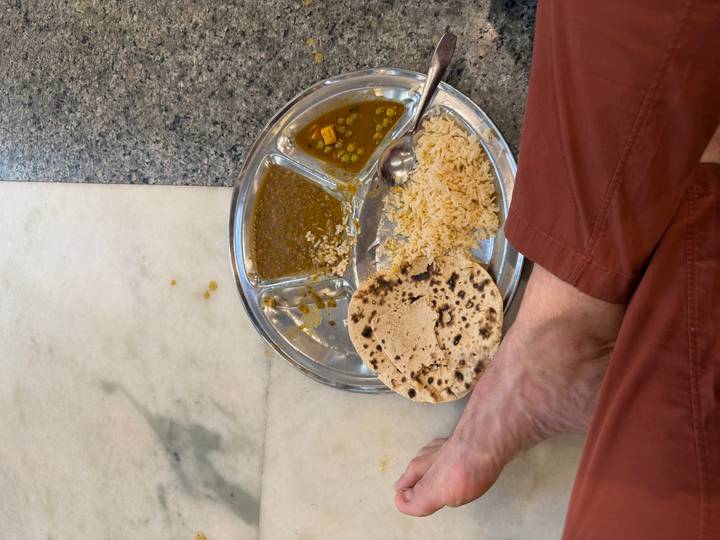 Simple Indian meal of dal, rice and chapati served on a steel tray on the floor with a person’s bare foot visible.