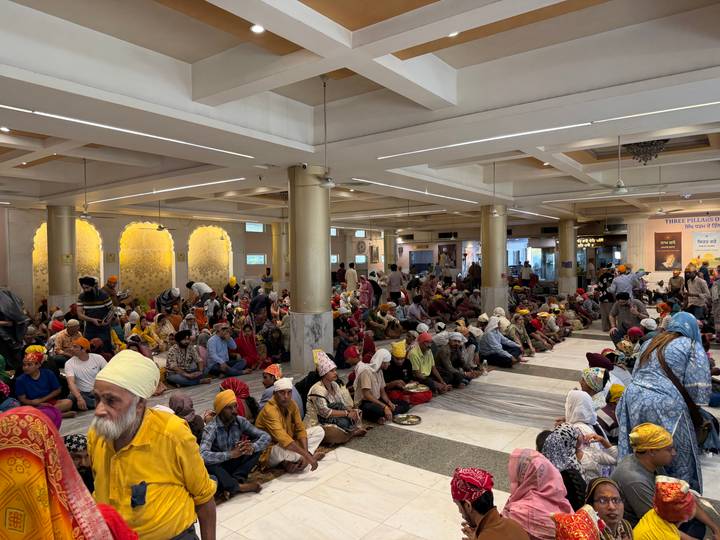 Crowded langar hall where hundreds of devotees sit on the floor awaiting a communal meal in a Sikh temple.