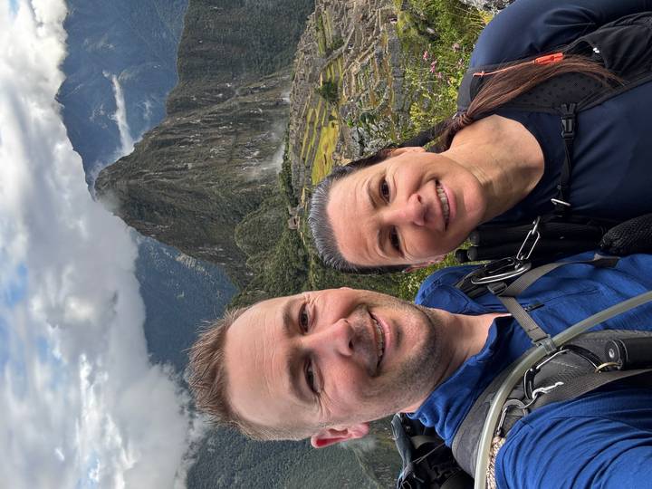 Smiling couple in trekking gear take a selfie with the iconic peak of Machu Picchu rising behind them under clouds.