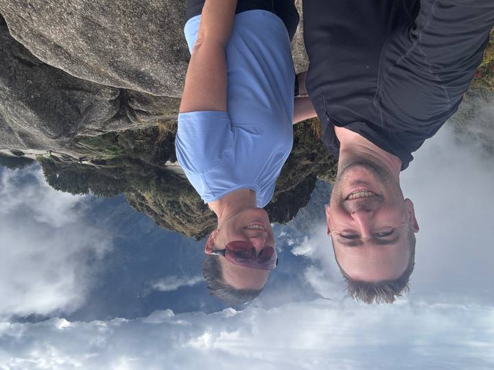 Two trekkers pose on a rocky ridge high in the Andes surrounded by clouds and dramatic peaks.