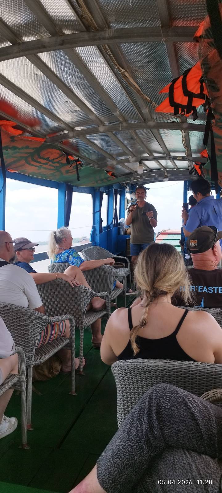 Travellers seated on wicker chairs aboard a river boat while a man sings with a microphone