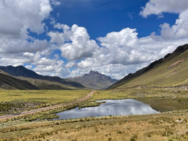 Wide Andean valley with railroad tracks, reflective pond and distant jagged peak beneath billowing clouds