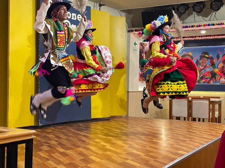 Traditional dancers in vibrant Andean costumes leap mid-air on a wooden stage indoors
