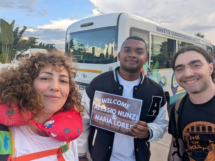 Smiling trio holds a welcome sign beside a shuttle bus.