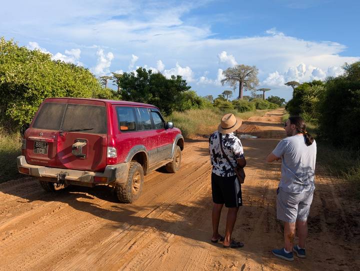Two men stand beside a muddy 4x4 on a red dirt road with distant baobab trees under blue sky.
