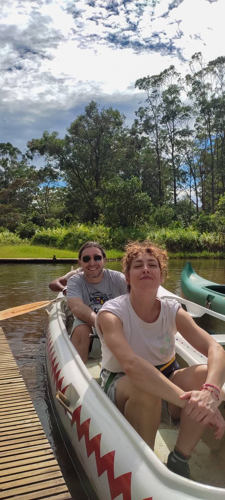Smiling couple paddle a canoe along a calm river bordered by lush greenery.