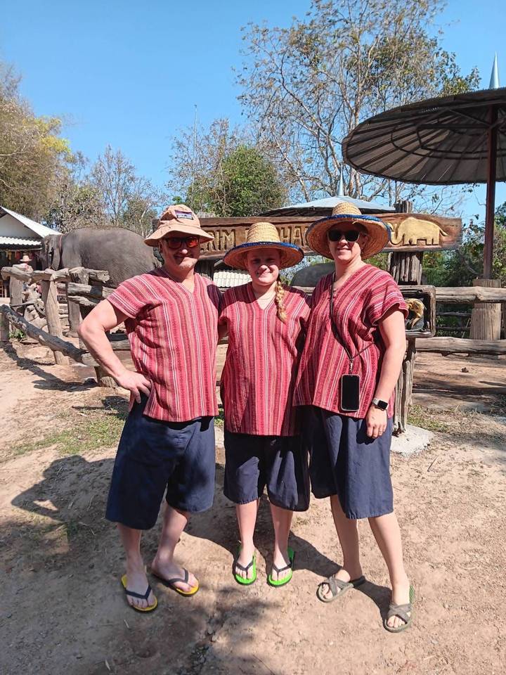 Travellers wearing traditional mahout outfits stand in front of an elephant camp enclosure.
