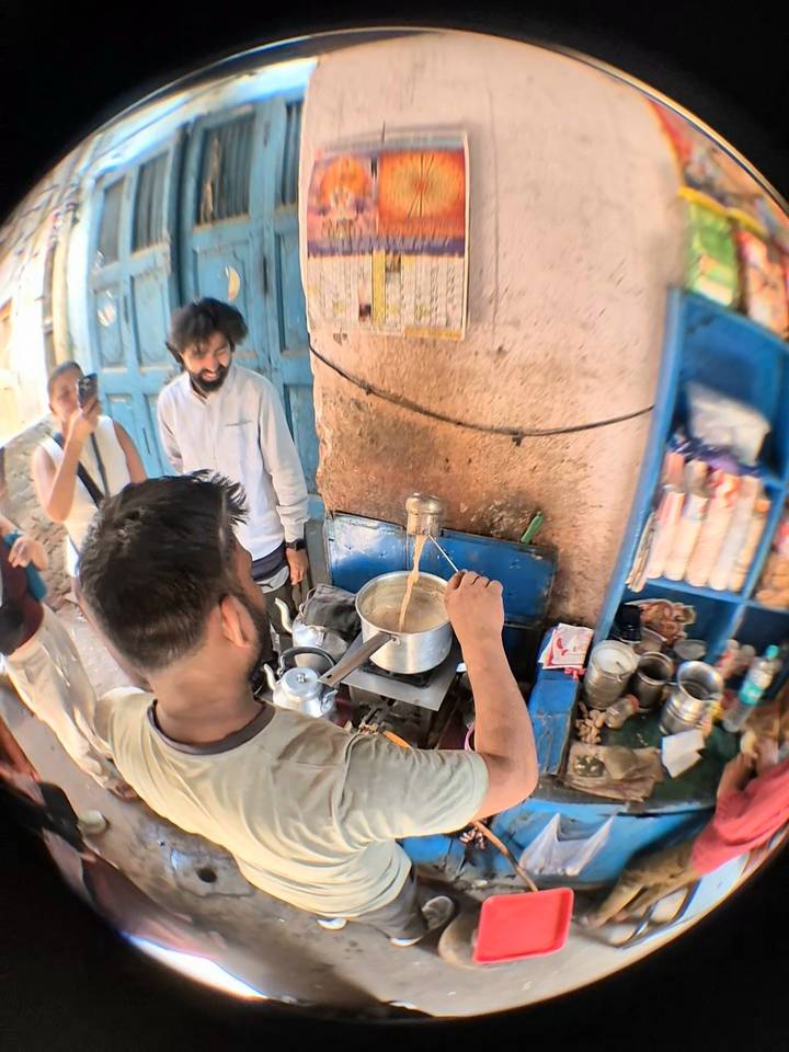 Street-side chai vendor pours tea into a pot while travellers watch inside a small, cluttered stall.