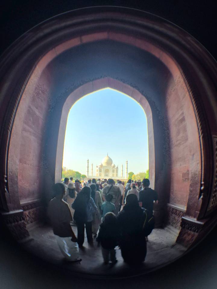 Crowds move through a grand red-sandstone arch framing a distant view of the Taj Mahal under a clear blue sky.