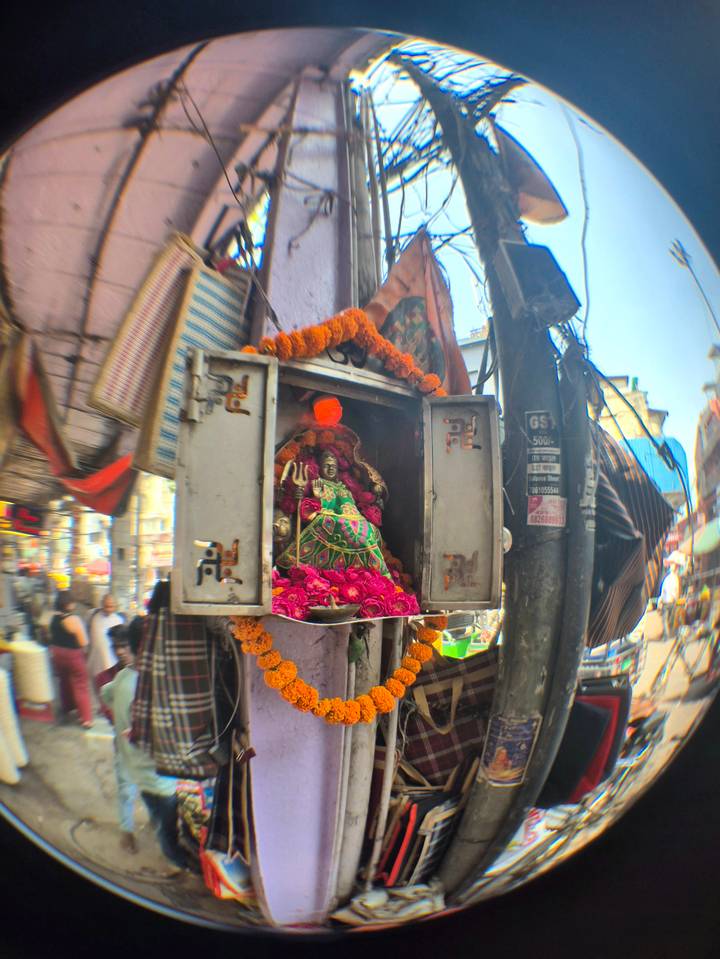 Small roadside Hindu shrine adorned with marigold garlands and bright pink roses amidst a bustling street.