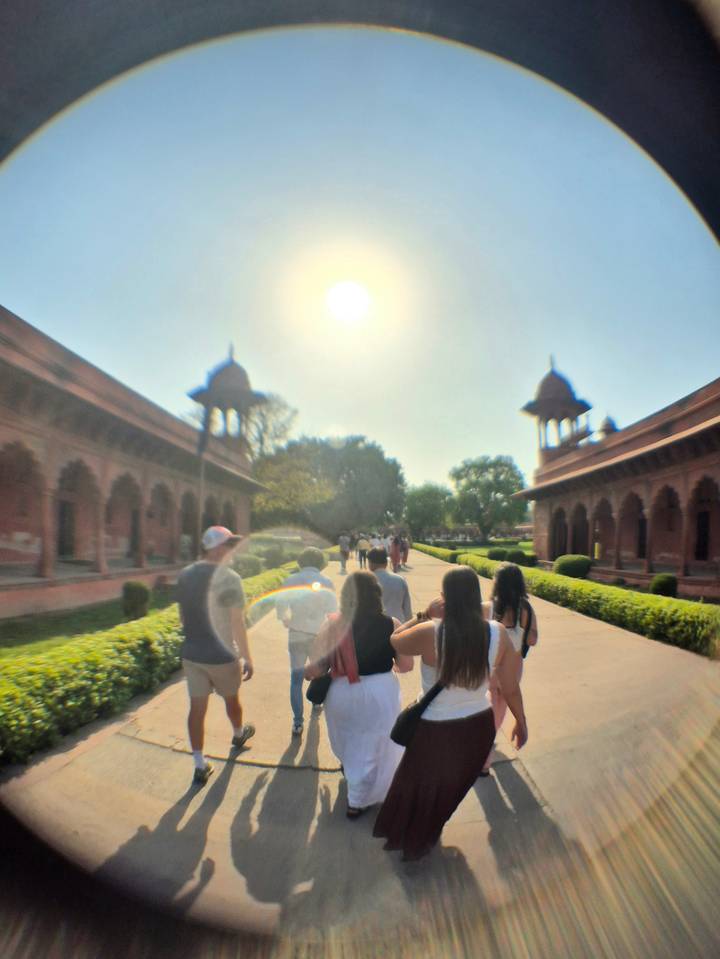 Tour group walking along a sun-drenched pathway between red sandstone buildings with bright midday sun overhead.