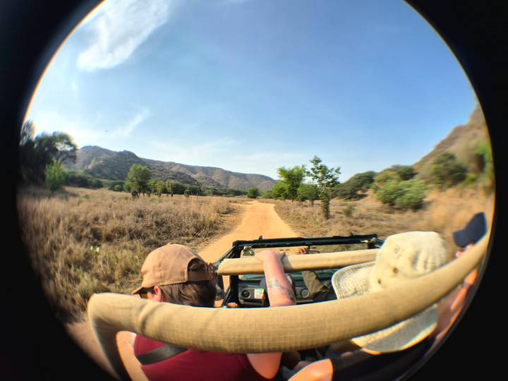 Travellers in an open jeep drive along a dusty safari track through dry scrub hills.