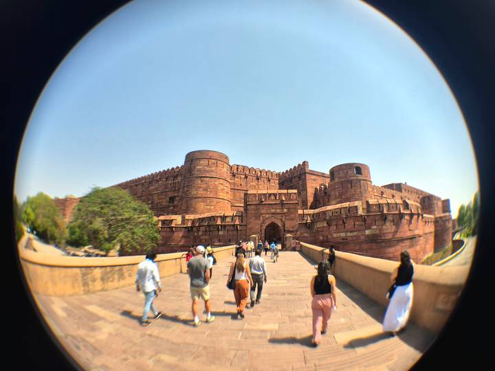 Visitors walk across a bridge toward the massive red-sandstone walls of Agra Fort under a clear sky.