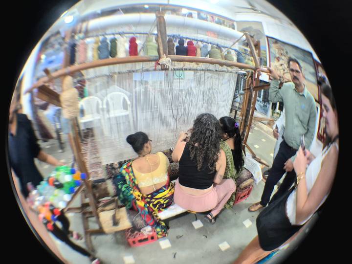Women watch artisans weave colourful carpets on a traditional loom inside a workshop.