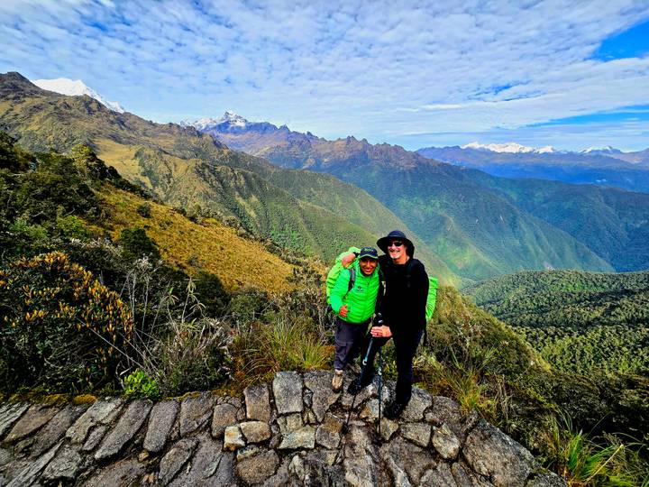 Two trekkers in bright gear smiling on a high ridge overlooking vast green ranges