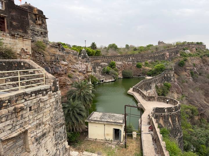 Stone fortifications snake along a rocky cliffside overlooking a narrow green reservoir
