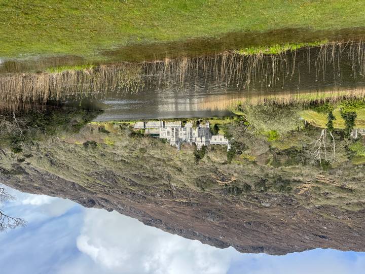 Historic grey-stone castle nestled beside a tranquil lake with wooded hills rising behind.