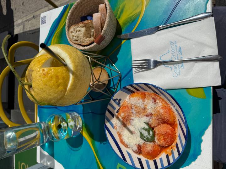 Close-up of Italian meal with gnocchi in tomato sauce, grated cheese and a refreshing drink served in a hollowed lemon on a colorful table.