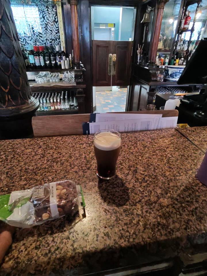 A pint of dark beer with creamy head sits on a granite bar counter in a cosy pub.