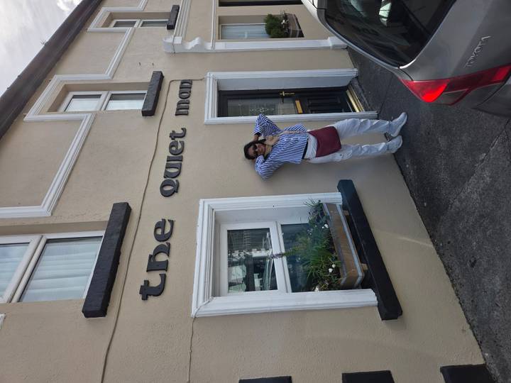Traveler posing in front of a beige building bearing the sign 'the quiet man'.