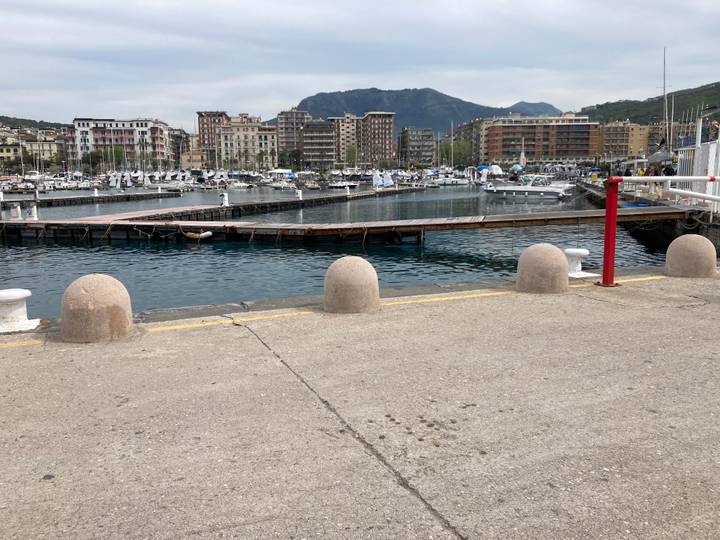 Marina with yachts and fishing boats docked beside a seaside town backed by low mountains under a cloudy sky.