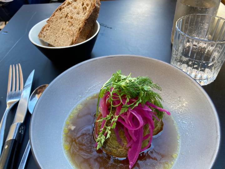 Bowl of braised dish garnished with bright pickled onions and fresh dill, served with rustic bread on a dark cafe table.