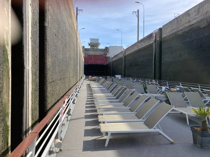 Cruise ship sun deck passing through a tall concrete lock chamber, rows of recliners stretching forward.