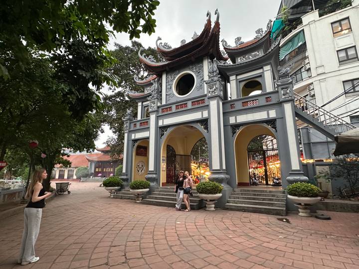 Historic pagoda-style entrance gate with ornate roof carvings, flanked by trees and visitors taking photos.