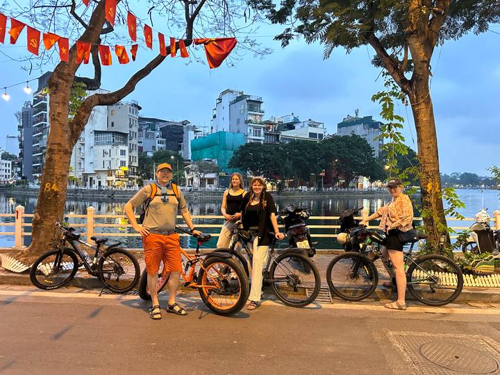 Group of cyclists posing with bikes beside an urban lake at dusk with city buildings in the background.