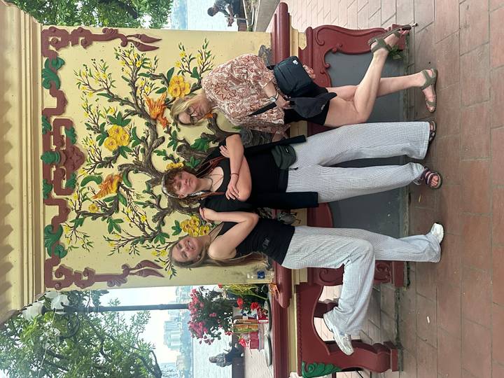 Three young women striking playful poses in front of an ornate wall mural with yellow blossoms.