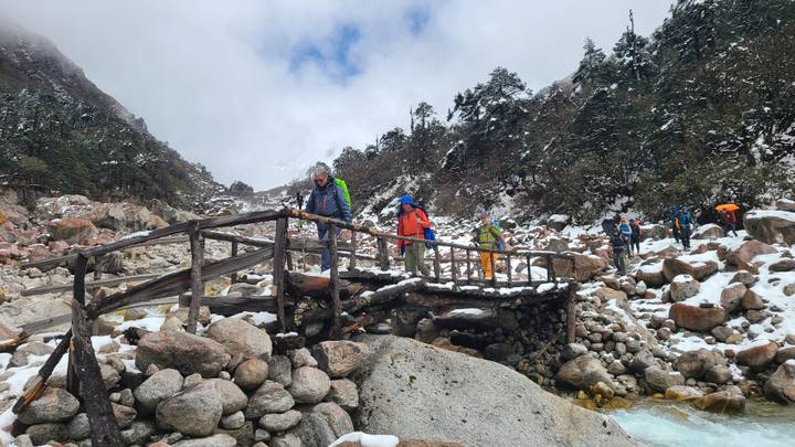 Hikers crossing a rustic wooden bridge over a snowy mountain stream