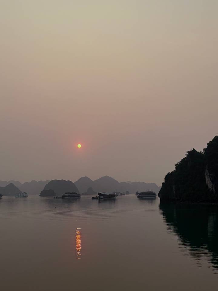 Hazy sunset over the karst islands of Ha Long Bay with cruise boats silhouetted on calm waters.