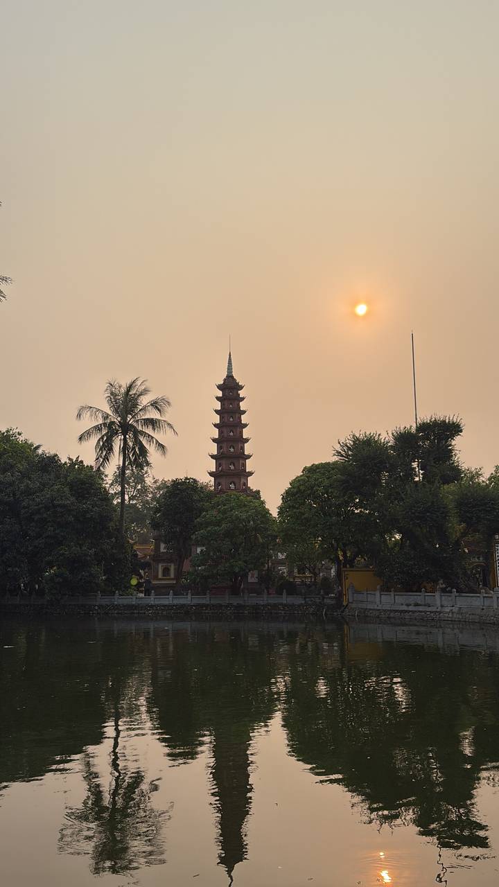 Tran Quoc Pagoda rises among trees beside West Lake under a hazy sunset sky in Hanoi.