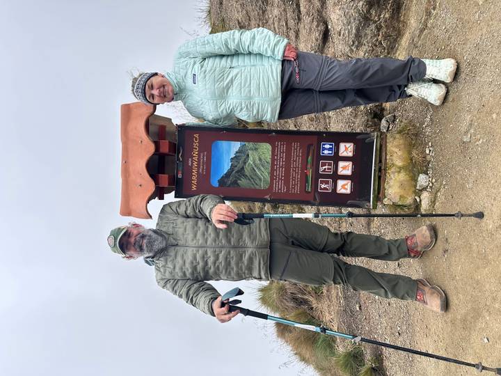 Two trekkers pose beside the Warmiwañusca Pass sign with misty peaks rising behind them on the Inca Trail.
