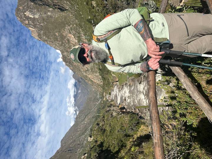 Bearded hiker with trekking poles stands beside a rushing Andean river against a backdrop of rugged mountains and blue sky.