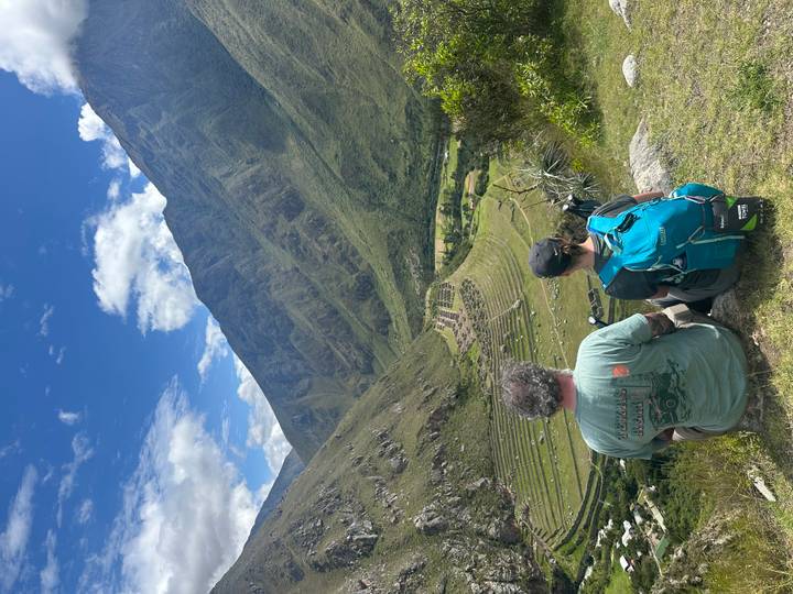 Pair of trekkers sit on a ridge overlooking ancient agricultural terraces and a verdant valley under a brilliant blue sky.