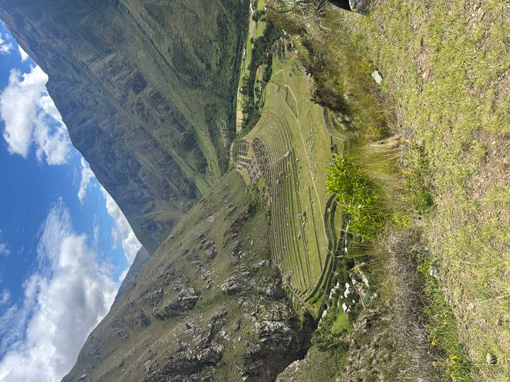 Wide view of the Patallacta ruins and surrounding Andean valley beneath towering green ridges and blue skies.