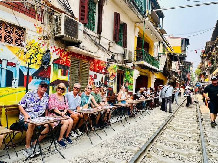 Tour group seated at tiny street-side tables beside Hanoi’s famous railway track, colorful murals and colonial facades behind them.