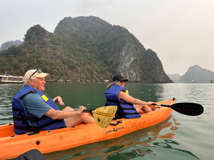 Two travelers paddle an orange kayak across calm jade waters surrounded by towering forested limestone cliffs in Halong Bay.